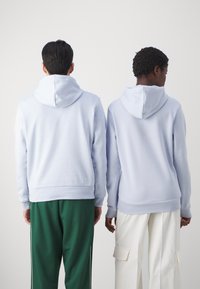 Moletons de capuz azul claro exibidos em duas pessoas, apresentando um corte relaxado e punhos canelados, combinados com calças verdes e brancas.