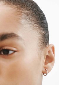 Pequeño pendiente de aro en metal de oro rosa con una superficie texturizada y una fila de piedras transparentes incrustadas. El diseño es simple y elegante.