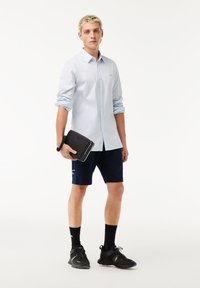 Light blue striped long-sleeve shirt, navy shorts, black sneakers, holding a black electronic case. Simple and contemporary casual wear.