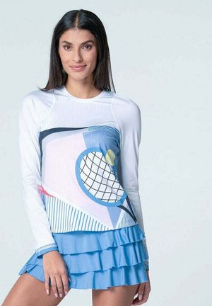 Frau in einem weißen Langarmoberteil mit einem Tennisschlägerdesign und einem mehrlagigen blauen Tennisrock vor einem hellen Hintergrund.