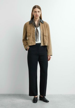 Woman wearing a tan jacket with leather collar, patterned scarf, white shirt, black trousers, and black shoes standing against a plain light wall.