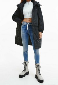 Abrigo de plumas negro con capucha y cierre en la parte delantera, combinado con una blusa blanca corta y jeans ajustados azules. Botas de combate grises con cordones completan el look.