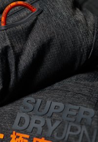 Tela gris oscuro con un patrón texturizado, que cuenta con un lazo naranja y un logo impreso en negro que dice "SUPER DRYJPN" en letras grandes.