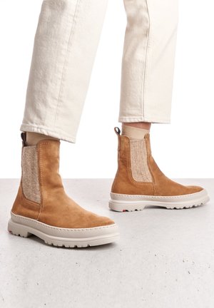 Persoon draagt beige suède enkellaarsjes met lichte stoffen inzetstukken en witte dikke zolen, gecombineerd met crèmekleurige broek.