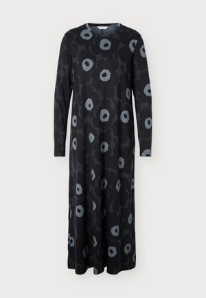 Femme portant une longue robe noire avec des motifs floraux gris, des manches longues et des chaussures noires à bout pointu, debout devant un fond uni.