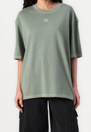 Person trägt ein lockeres, verblasstes grünes Adidas-T-Shirt mit kurzen Ärmeln und schwarze Hosen mit weitem Bein und genähten vertikalen Linien.