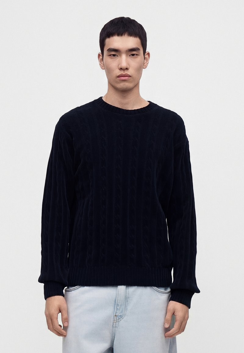 Jeune homme aux cheveux courts et foncés portant un pull en maille torsadée noir et un jean bleu clair, debout devant un fond blanc uni.