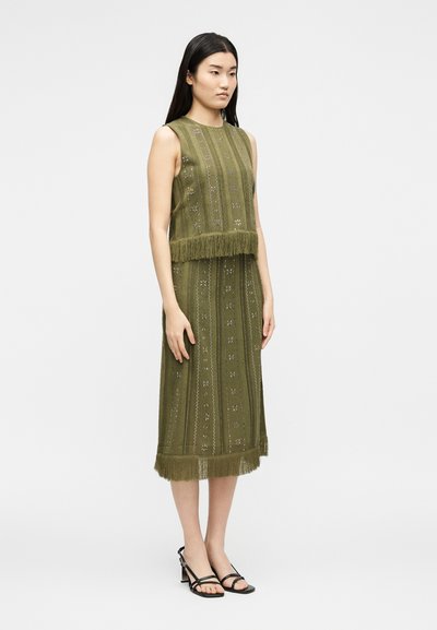 Γυναίκα που φοράει ελιά πράσινο αμάνικο φόρεμα με κρόσσια και μοτίβο υφής, στέκεται με τα χέρια χαλαρά στο πλάι, μαύρα σανδάλια με τακούνι.