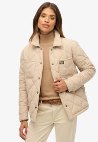 Beige gesteppte Jacke mit Kragen, vorderem Knopfverschluss und Brusttasche. Getragen über einem hellbraunen Rollkragenpullover, kombiniert mit beigen Hosen.