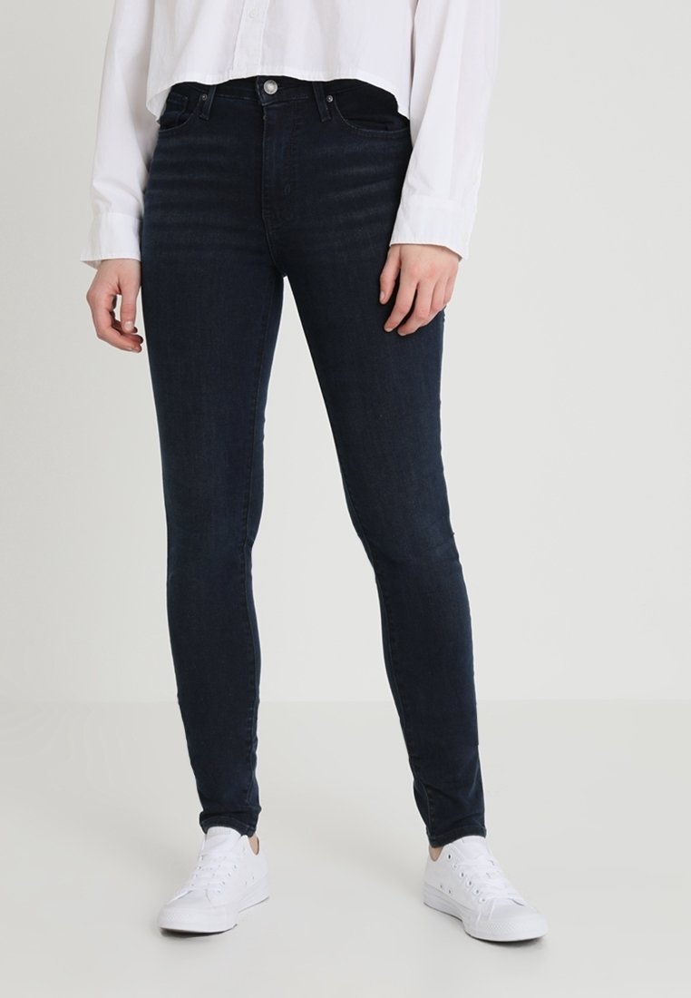 Tmavé džíny skinny s vysokým pasem, který mají klasický pětikapsový design a jemné prošívání, doplněné bílými teniskami.