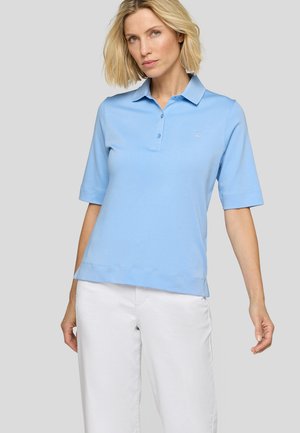Frau mit kurzen blonden Haaren, die ein hellblaues kurzärmeliges Poloshirt und weiße Hose trägt, steht vor einem schlichten grauen Hintergrund.
