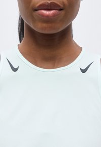 Κοντινό πλάνο ενός ατόμου που φοράει ανοιχτό μπλε αμάνικο τοπ με μαύρα λογότυπα Nike κοντά στους ώμους, σε απλό λευκό φόντο.