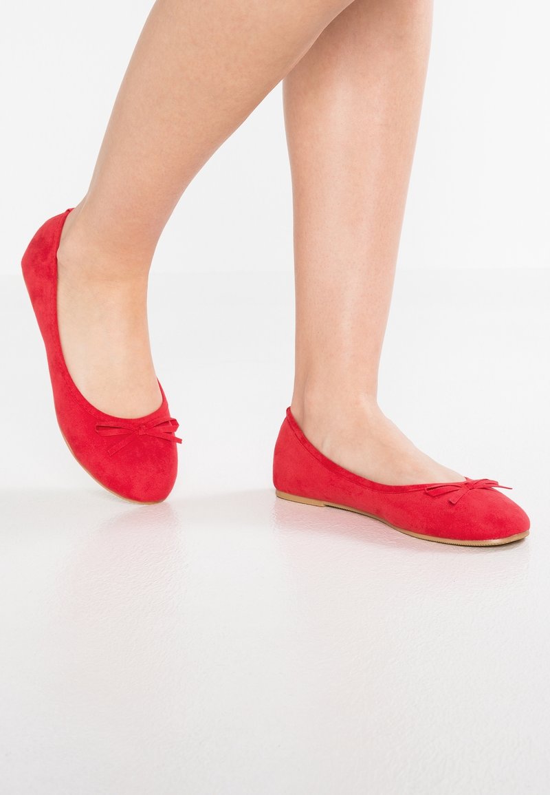 Jambes portant des ballerines rouges vives avec de petits nœuds sur un sol blanc lisse.