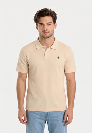 Joven con cabello corto y castaño, vestido con una camiseta tipo polo beige de manga corta y jeans azul claro, posando frente a un fondo blanco liso.