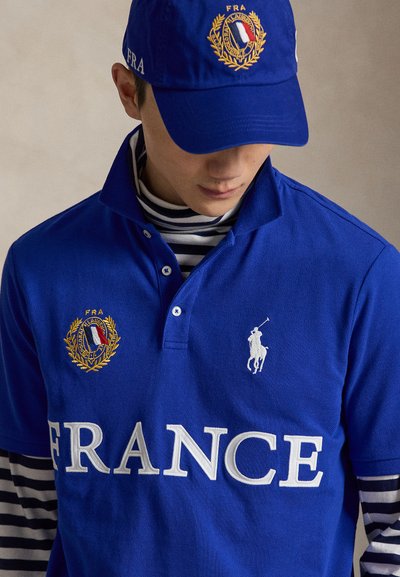 Hombre con gorra azul y polo con emblema de Francia, logo blanco de jugador de polo y gran texto "FRANCE", con camisa a rayas debajo.