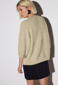 Hellgoldener Pullover mit weicher Textur und dreiviertel Ärmeln, kombiniert mit einem dunklen karierten Rock. Das Model posiert von hinten und präsentiert das Kleidungsstück.