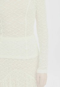 Top en dentelle de couleur crème clair, avec des manches longues et un corsage ajusté orné d'un motif floral et d'une finition texturée. Détails avec une ceinture lisse.