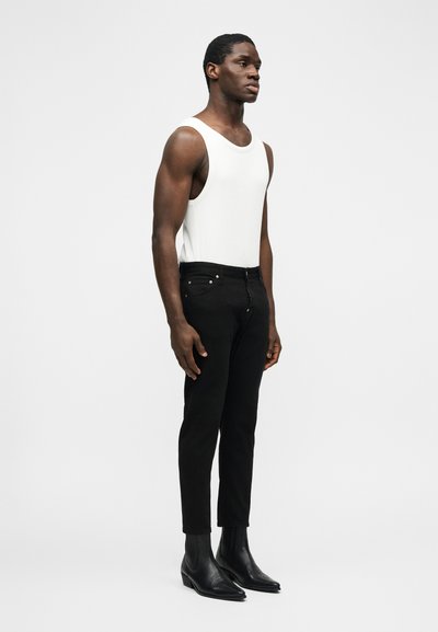Homme debout portant un haut sans manches blanc, un pantalon noir slim et des bottines noires à bout pointu sur un fond uni.