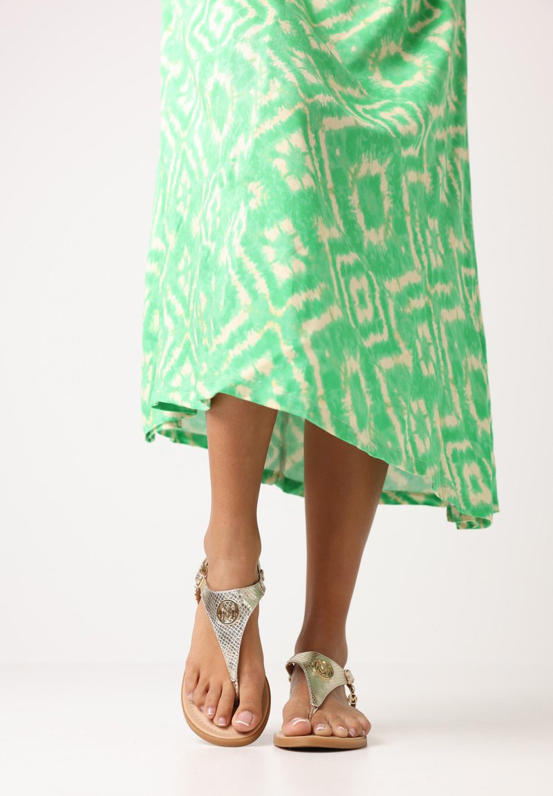 Metalen zilveren sandalen met een T-band ontwerp, voorzien van gedetailleerde hardware. De getoonde voet wordt aangevuld door een levendig groen patroonjurk.