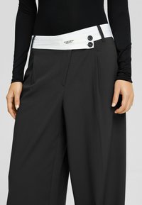 Svarta vida byxor med en kontrasterande vit midja som har två knappar. Tyget ser slätt ut med en strukturerad passform och sidofickor.