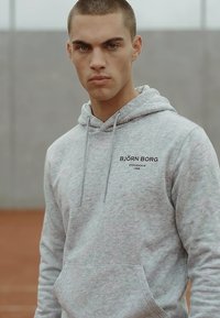 Mladý muž s krátkymi vlasmi v svetlosivom pulóvri s logom "BJÖRN BORG", stojaci vonku na rozmazanom sivom a hnedom pozadí.