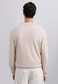 Pull en polo beige clair à manches longues avec col et poignets côtelés, texture lisse, design ajusté et détails de couture minimaux.