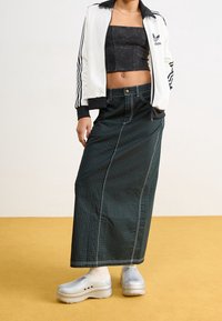 Crop top noir, veste de survêtement blanche avec des rayures noires, jupe à rayures vertes, chaussures à plateforme grises, debout sur un sol jaune.