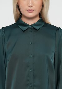 Mørkegrønn satengbluse med klassisk krage og knappelukking foran, med lange ermer og myk, glatt tekstur.