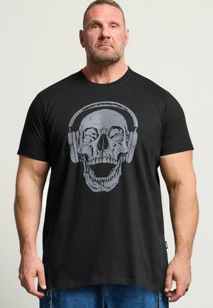 Mężczyzna w czarnej koszulce z dużą szarą czaszką w słuchawkach nadrukowaną z przodu, stojący na jednolitym jasnym tle.