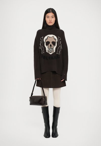 Μαύρος πλεκτός πλούνα με γραφικό κρανίο και κείμενο "VOLTAIRE", συνδυασμένος με καφέ φούστα, λευκές καλσόν και μαύρες μπότες μέχρι το γόνατο. Καφέ τσάντα.