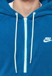 Uomo che indossa una felpa con cappuccio in pile blu Nike, con cordini bianchi e cerniera azzurro chiaro, visibile collo e parte inferiore del viso.