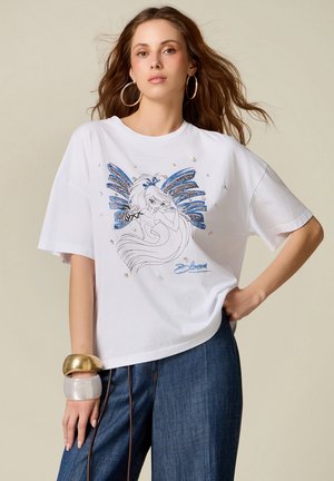 Jonge vrouw met los haar die een wit T-shirt draagt met een afbeelding van een blauw gevleugelde fee en donkerblauwe spijkerbroek, voorzien van grote armbanden.