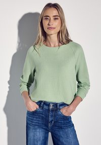 Ljusgrön ribbad tröja med långa ärmar, rund halsringning, kombinerad med mörkblå denimjeans. Modellen står med händerna i fickorna.
