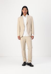 Giacca da abito beige con pantaloni abbinati, davanti abbottonato, tasche piatte, abbinata a una camicia bianca e scarpe nere, su uno sfondo semplice.