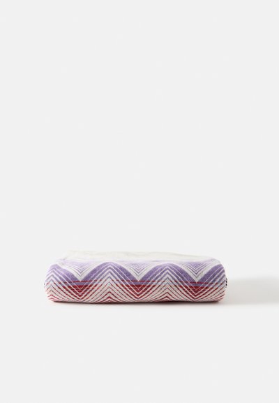 Couverture tissée pliée avec motifs chevrons en violet, rouge et blanc sur un fond blanc uni.