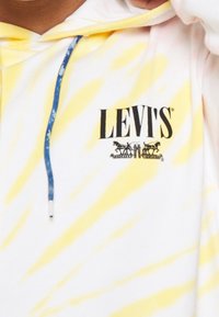Felpa bianca con motivo tie-dye giallo, dotata di cordino blu e logo "LEVI'S" stampato in nero con cavalli sopra. Texture morbida.