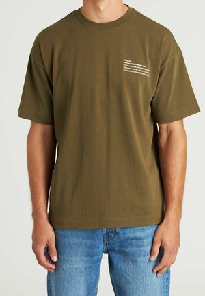 Hombre con camiseta de manga corta verde oliva con texto pequeño blanco en el pecho izquierdo y jeans azules, de pie frente a un fondo neutro.