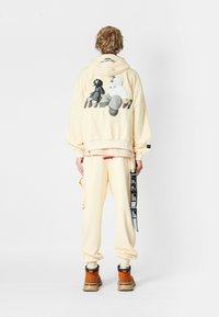 Joven con cabello rizado que lleva puesta una sudadera beige y pantalones a juego con detalles gráficos, y botas marrones, de pie frente a un fondo liso.