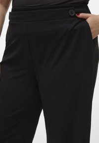 Pantalons taillés noirs avec une texture lisse, dotés d'un bouton à l'avant, de plis et de poches latérales pour un usage pratique.