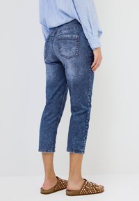 Džínové ankle jeans v tmavém odstínu s opotřebovaným vzhledem, pěti kapsami a rovnými nohavicemi. V kombinaci s hnědými sandály na platformě s puntíky.