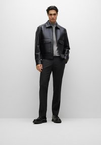 Giubbotto di pelle nera con due grandi tasche frontali, maglione grigio con zip, pantaloni neri su misura e scarpe nere lucide. Texture liscia in tutto.