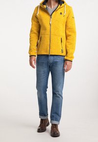 Gul marl zip-up huvtröja med marinblå accenter, två framfickor med dragkedja och dragsnören. Kombinerad med upprullade blå jeans och bruna stövlar.