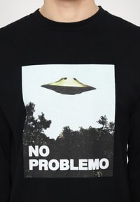 Black long-sleeve shirt featuring a printed graphic of a UFO above trees with the text "NO PROBLEMO" in bold white letters.