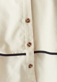 Primo piano di una camicia di stoffa crema con tre bottoni marroni e una striscia nera orizzontale sotto i bottoni.