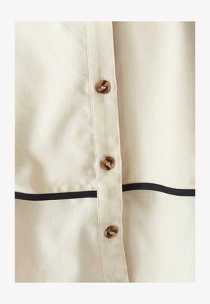 Close-up of cream fabric shirt front with three brown buttons and a horizontal black stripe below the buttons.
