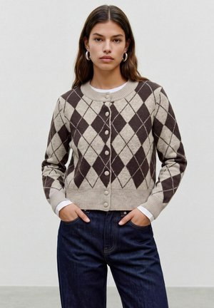Femme aux longs cheveux foncés portant des boucles d'oreilles créoles argentées, un cardigan beige et marron à motif losange boutonné, et un jean bleu foncé, debout les mains dans les poches.