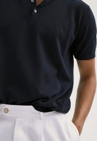 Homme portant un polo noir à manches courtes, rentré dans un pantalon blanc avec fermeture par boutons, une main dans la poche.