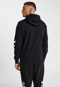 Svart hoodie med huva och ribbade ärmar, har vita diagonala ränder på vänster ärm och en avslappnad passform. Bärs med svarta byxor.