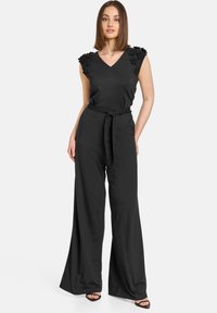 Schwarzer Jumpsuit mit V-Ausschnitt und verzierten Kappenärmeln, ausgestattet mit einem Taillengürtel und weitem Bein. Glattes Material und lässige Passform.