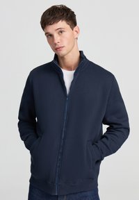 Felpa blu navy con zip, realizzata in materiale morbido. Presenta un colletto alto, maniche lunghe e tasche laterali; texture liscia con polsini a coste.